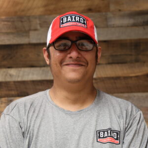 Headshot of co-foreman, Julian Munoz, taken at the San Antonio office. He is smiling and wearing a light grey Baird t-shirt and red and white Baird baseball cap.