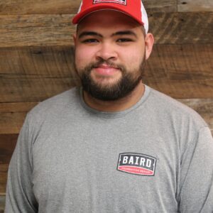Headshot of installer, John Gonzalez, taken at the San Antonio office. He is wearing a gray long-sleeve Baird t-shirt and a red and white Baird baseball cap.