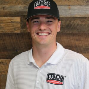 Headshot of commercial solutions specialist, Will Chaney, taken at the San Antonio office. He is smiling and wearing a white with thin black horizontal stripes Baird polo shirt and a black Baird cap.