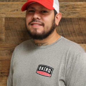 Headshot of installer, Derick Marin, taken at the San Antonio office. He is smiling and wearing a red and white Baird baseball cap and gray long sleeve Baird t-shirt.
