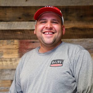 Headshot of installer, Albert Sierra, taken at the San Antonio office. He is smiling and wearing a red and white Baird cap and gray long-sleeve Baird t-shirt.