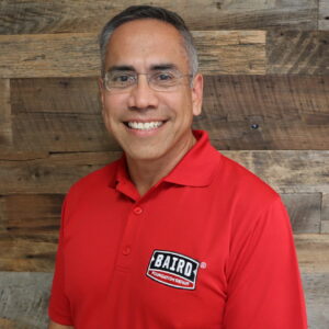 Headshot of system design specialist, Rey Rodriguez, taken at the Corpus Christi office. He is smiling, wearing rimless glasses, and wearing a red Baird Foundation Repair polo shirt.