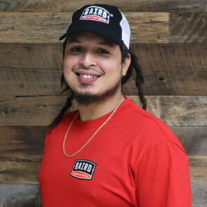 Headshot of installer, Shawn Ladson, taken at the San Antonio office. He is smiling and wearing a red Baird t-shirt, a gold chain necklace, and a black and white Baird baseball cap.