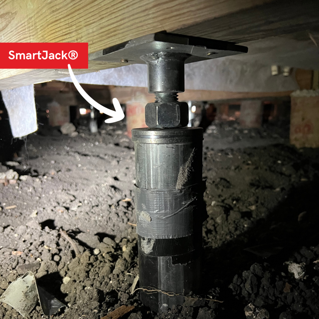 Close up of a steel SmartJack installed in the crawlspace underneath wooden foundation beams with a SmartJack logo and white arrow graphic pointing to the SmartJack.