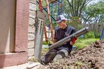 Technician installing a steel foundation support pier beside a home to stabilize and repair structural settling.
